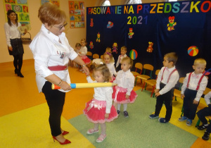 Pani dyrektor Maria Królikowska trzyma w prawej dłoni dużą żółtą kredką, którą przykłada dziewczynce do lewego ramienia. W lewej dłoni trzyma dłoń dziewczynki. W tle stoją dzieci i Pani Ewelina.